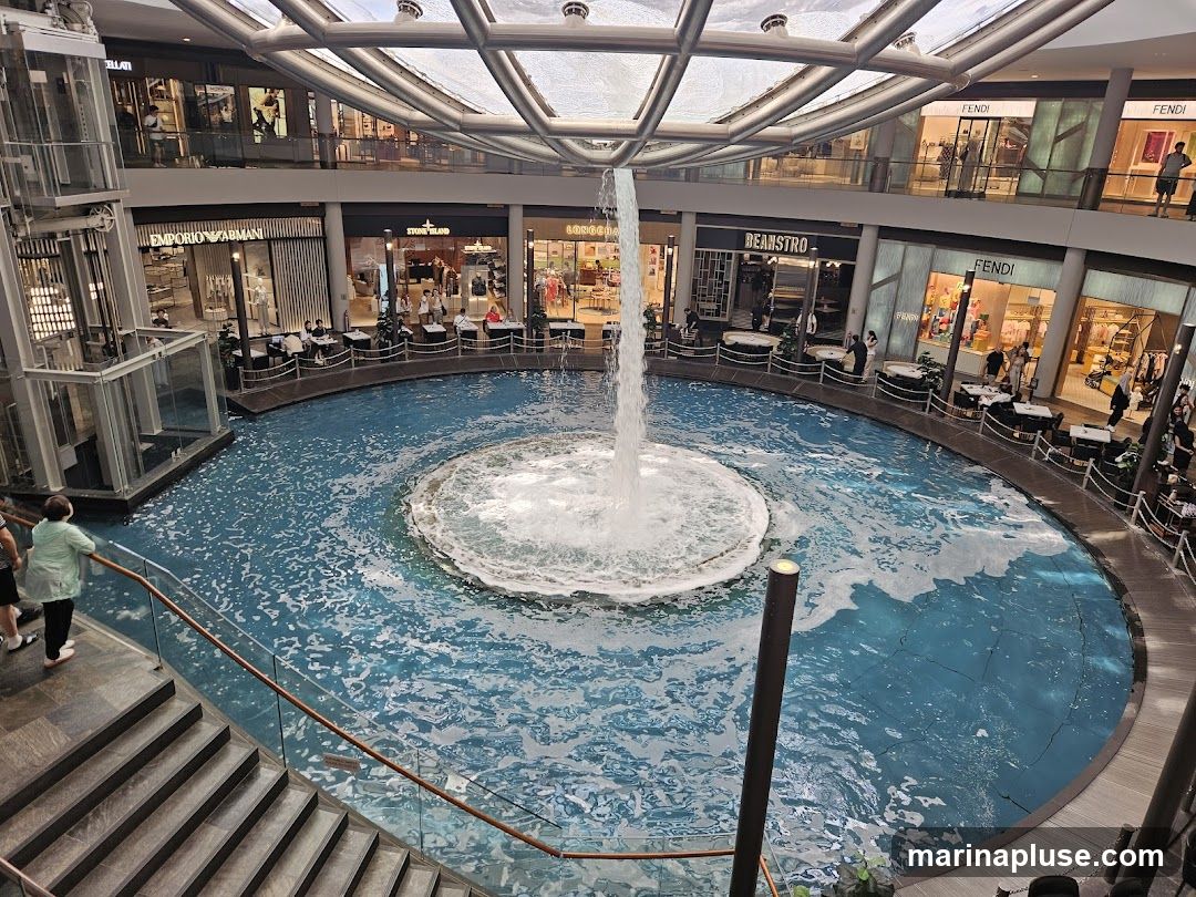 An indoor shopping mall features a large, circular water feature with a fountain in the center. Surrounding the water feature are multiple levels of shops and restaurants. Escalators and elevators are visible, along with seating areas around the water. People are seen walking, dining, and shopping. Brands like Emporio Armani, Stone Island, Beanstro, and Fendi are visible on the storefronts. The mall has a modern design with a glass and metal ceiling structure.