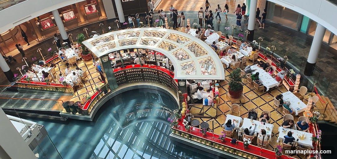 An aerial view of a busy indoor dining area within a shopping mall. A curved bar with display cases is centrally located, surrounded by tables where patrons are seated and dining. The floor is decorated with a geometric pattern. In the background, other shops and shoppers are visible.