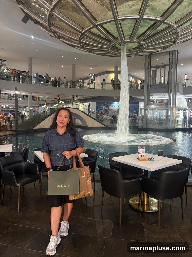 A woman stands in a modern shopping mall, holding shopping bags. Behind her, a large indoor fountain with a cascading water feature is visible. The mall has multiple levels with shops and people browsing. The woman is wearing a denim shirt and a black skirt, with athletic shoes. She is carrying a green Longchamp shopping bag and a patterned tote bag. In the foreground, there are tables and chairs, suggesting a dining or seating area. A small sign on one of the tables indicates it is reserved.