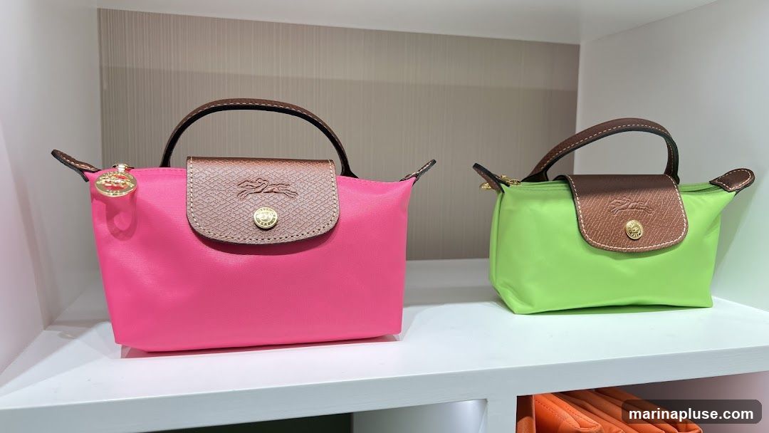 A bright pink and a lime green cosmetic bag are displayed side-by-side on a white shelf. Both bags feature brown leather trim and handles, and a gold-toned emblem with a horse and rider. The pink bag has a gold charm attached to its zipper pull.