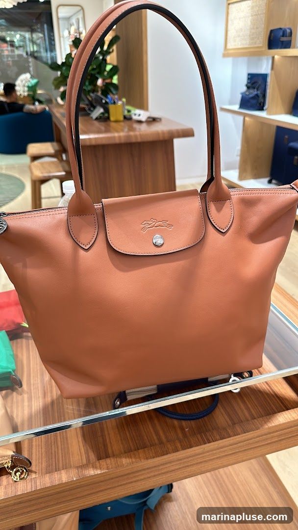 A close-up shot of a brown leather tote bag with the Longchamp logo embossed on the flap. The bag is displayed on a wooden surface with a glass top, likely in a retail store. In the blurred background, a person is seated on a blue chair, and shelves display other bags and accessories.