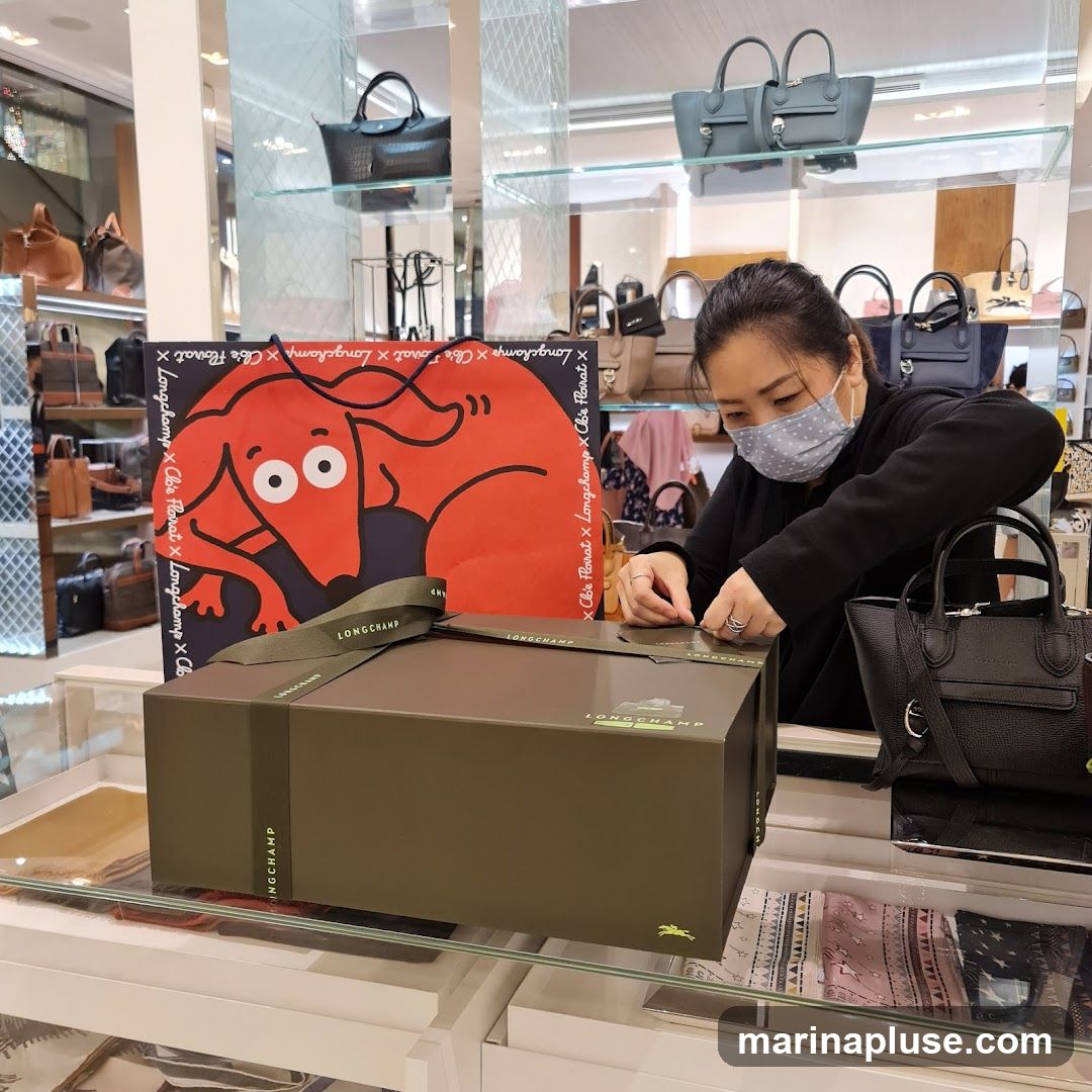 A woman wearing a face mask is wrapping a gift box with ribbon. The box and ribbon have the Longchamp logo. Behind her, shelves are stocked with various handbags. A large red shopping bag with a cartoon dog illustration and Longchamp branding is also visible.
