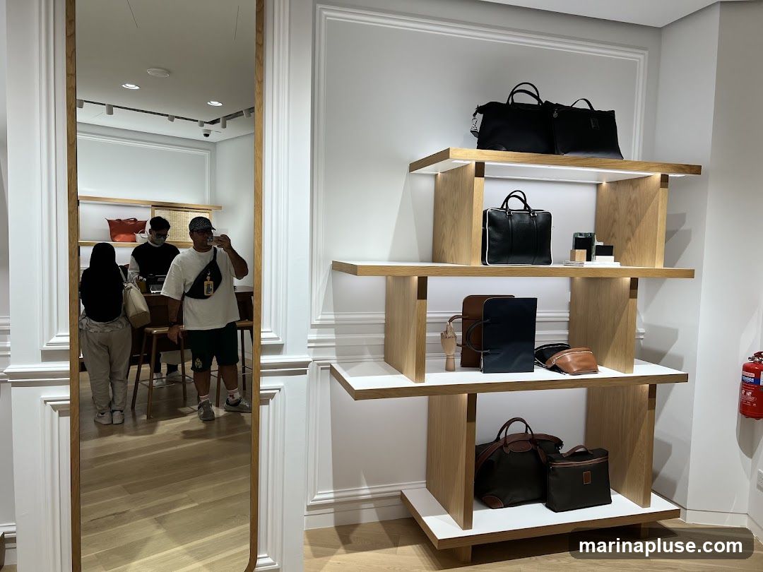 A well-lit store interior displays various handbags on wooden shelves. In the foreground, a large wooden shelf unit holds several bags, including black briefcases, brown and navy folders, and a tan crossbody bag. The lower shelf features a large dark duffel bag and a smaller dark cosmetic bag. A mirror on the left reflects the store's interior, showing customers and staff. A fire extinguisher is visible on the right wall.