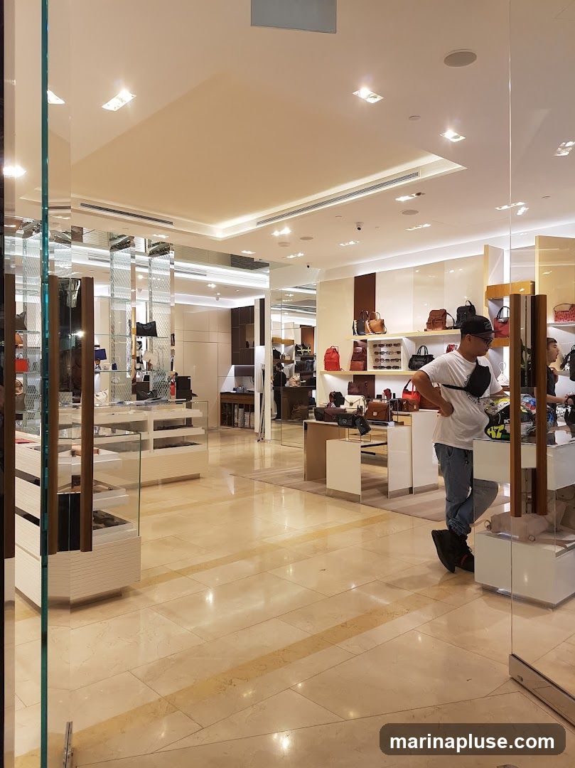 A view from outside a store, looking into a well-lit interior displaying a variety of handbags and accessories on shelves and display cases. Several customers are browsing the merchandise. The flooring is polished marble.