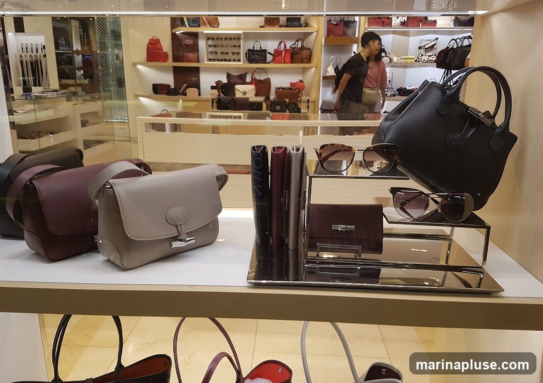 A display of various handbags, sunglasses, and wallets on shelves and stands within a store. The handbags are in different colors and styles, including a burgundy shoulder bag and a light beige crossbody bag in the foreground. Sunglasses are displayed on tiered stands, and a dark brown wallet is also visible. In the background, more handbags and accessories are arranged on shelves, and two people are browsing in the store.