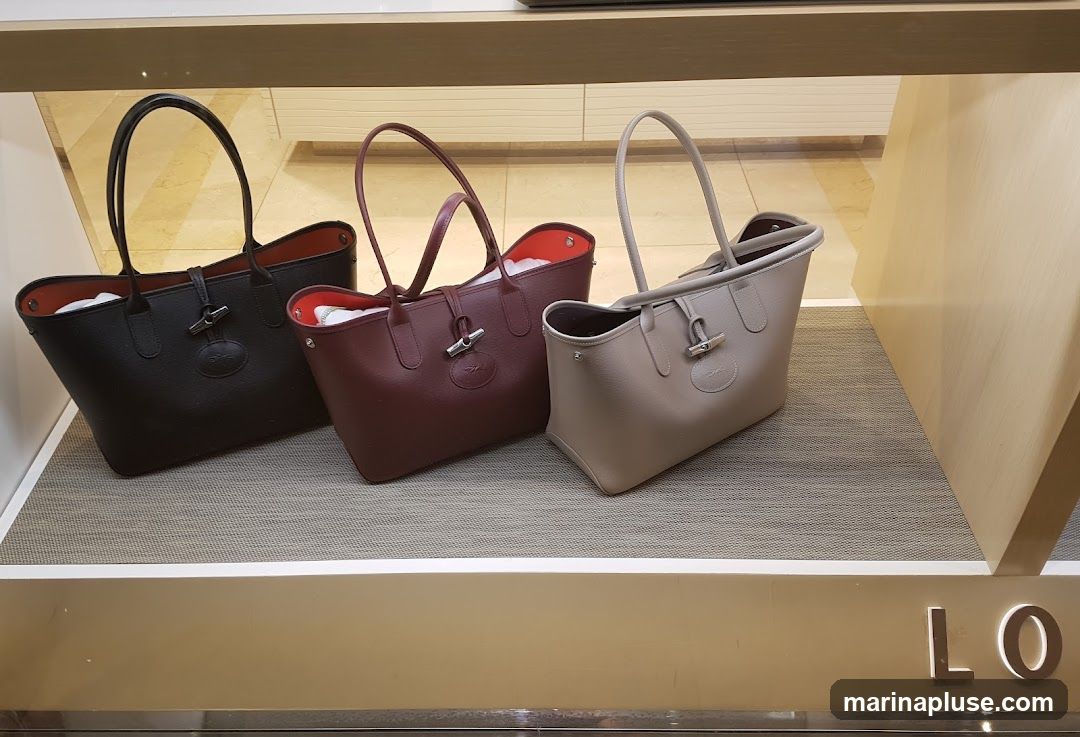 Three leather tote bags are displayed on a shelf. The bags are in black, burgundy, and beige. Each bag has a textured finish and a silver clasp closure. The interior of the bags appears to be a contrasting color. The bags are arranged in a row, with the black bag on the left, the burgundy bag in the middle, and the beige bag on the right. The display shelf is lit from above, and the background appears to be a store interior. Part of a store sign with the letters "LO" is visible at the bottom right.
