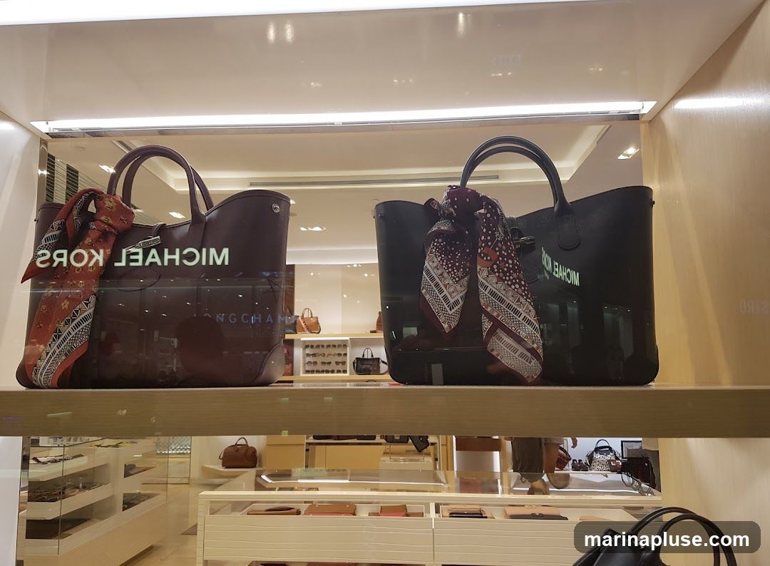 A display window showcases two large designer handbags, one burgundy and one black, each adorned with a patterned scarf tied to the handle. The burgundy bag has the brand name "MICHAEL KORS" reflected on its surface. The black bag also has "MICHAEL KORS" visible. In the background, more handbags and accessories are visible on shelves, along with other store displays.