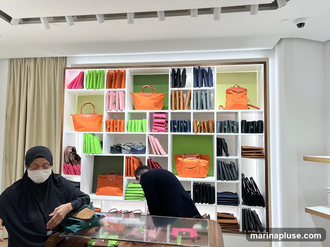 A retail store interior with a large display shelf filled with various handbags and accessories. The shelves are organized into cubbies, showcasing bags in bright colors like orange, pink, and green, as well as neutral tones like black, grey, and brown. Two customers are visible, one wearing a hijab and face mask, and another person in the background. The overall impression is a well-organized and brightly lit retail environment focused on handbags.