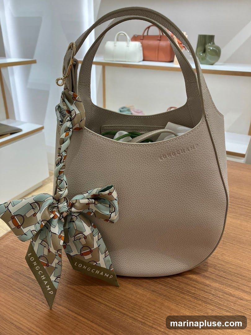 A light beige Longchamp handbag with a patterned scarf tied to the handle. The scarf has a print of horses and equestrian elements, with the Longchamp logo visible on the ends. The bag is textured and appears to be made of leather. In the background, shelves display other handbags and decorative items.