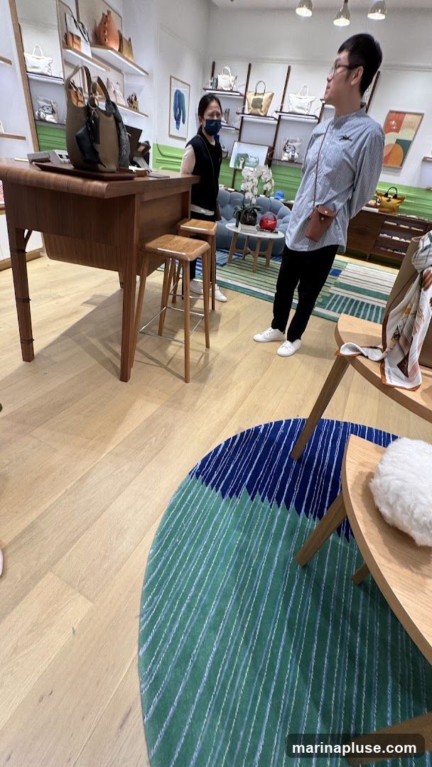 A retail store interior with shelves displaying various handbags. Two people are present, one standing behind a wooden counter and another looking towards the shelves. The store has a modern and clean aesthetic with wooden floors and minimalist decor. A colorful rug is visible on the floor.