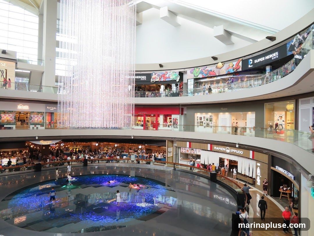 An interior view of a multi-level shopping mall featuring a large, circular water feature at the bottom level with people in it. Above the water feature, on the second level, are various shops and restaurants, including 'Ramen Ippudo' and 'Starbucks Coffee'. A large, sparkling crystal chandelier hangs from the ceiling in the center of the atrium. The upper levels also show storefronts and people browsing. The overall ambiance is bright and modern.