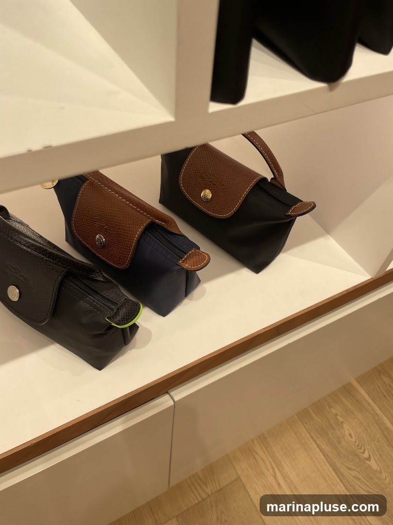 Three cosmetic bags are neatly arranged on a white shelf. The bags are made of nylon with leather trim and gold-tone hardware. One bag is black, another is navy blue, and the third is dark gray. The shelf is part of a larger display unit, with other items visible above and below. The flooring appears to be light-colored wood.