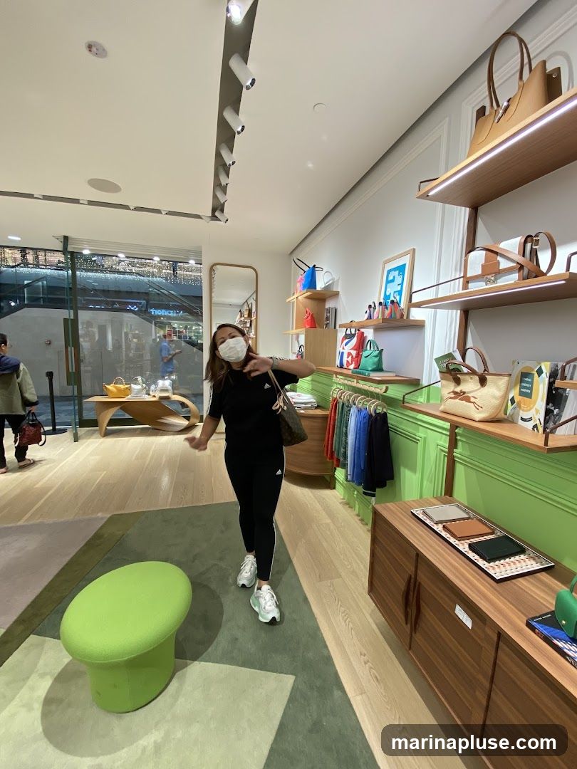 A woman wearing a face mask and athletic wear stands in a brightly lit store, posing for a photo. The store appears to be a high-end boutique selling handbags and accessories. Shelves display various bags, wallets, and clothing items. A lime green stool is in the foreground. In the background, through a glass partition, a shopping mall interior with escalators and other stores is visible.