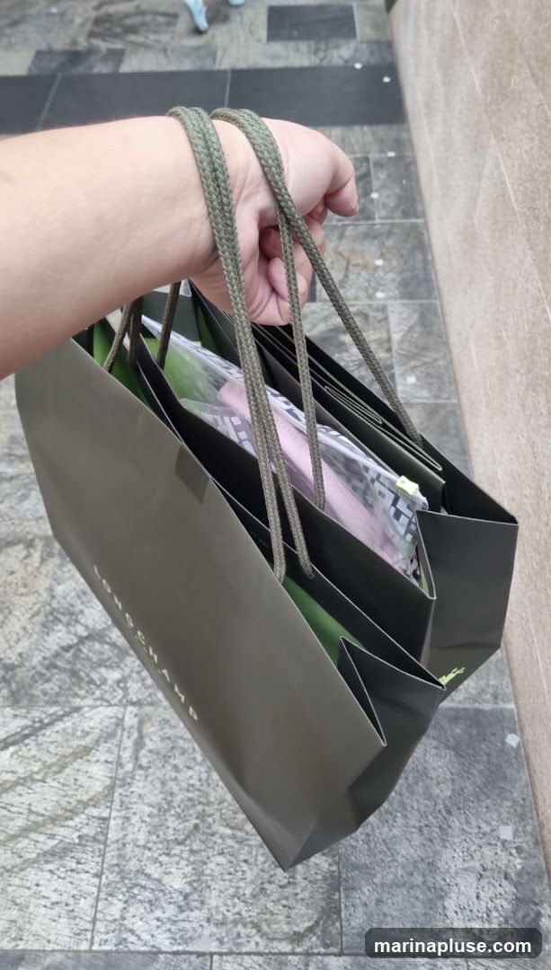 A hand holds several dark green shopping bags with rope handles. The bags have the word "LONGCHAMP" printed in gold lettering on the side. Items are visible inside the bags, including what appears to be pink clothing and clear plastic packaging. The bags are being carried outdoors on a tiled walkway.