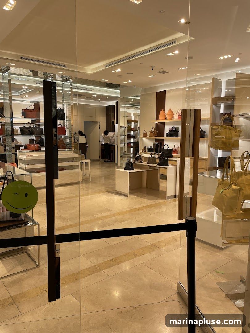 A view into a store selling handbags. The store has a clean, modern aesthetic with bright lighting. Shelves are stocked with various bags in different colors and styles. Some bags are displayed on mannequins or stands. There are also some decorative items on shelves. Two employees are visible in the background, one assisting a customer. A green smiley face sticker is visible on a display case near the entrance.