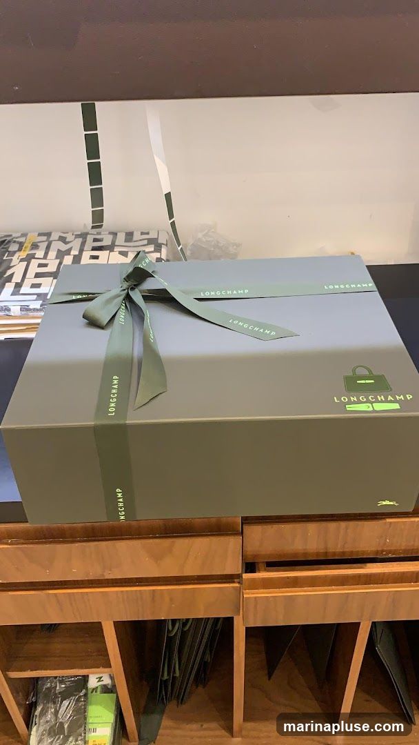 A large, grey gift box is tied with a green ribbon that has the Longchamp logo printed on it. The box also features a green Longchamp logo of a handbag and a horse. The box is placed on a wooden counter with shelves below, which contain various items, including what appear to be shopping bags.