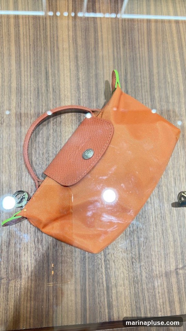 An orange pouch with a leather flap and snap closure is displayed on a wooden surface. The pouch has a small handle and a zipper closure. The surface appears to be a display case with reflections.
