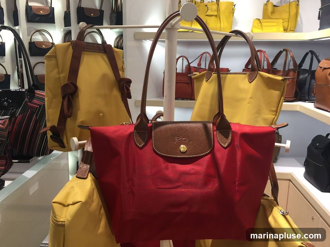 A collection of handbags in various colors and styles are displayed on shelves and racks. The foreground features a red nylon tote bag with brown leather trim and handles. Behind it, a yellow backpack with brown straps is visible. Further back, more handbags in yellow, red, and black are arranged on shelves. The overall impression is of a retail store display for fashion accessories.
