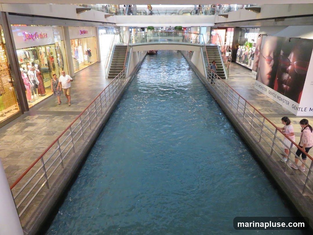 An indoor mall features a canal running through its center, with shops and walkways on either side. People are seen walking along the pathways and looking at the shops. The architecture includes escalators and bridges spanning the canal.
