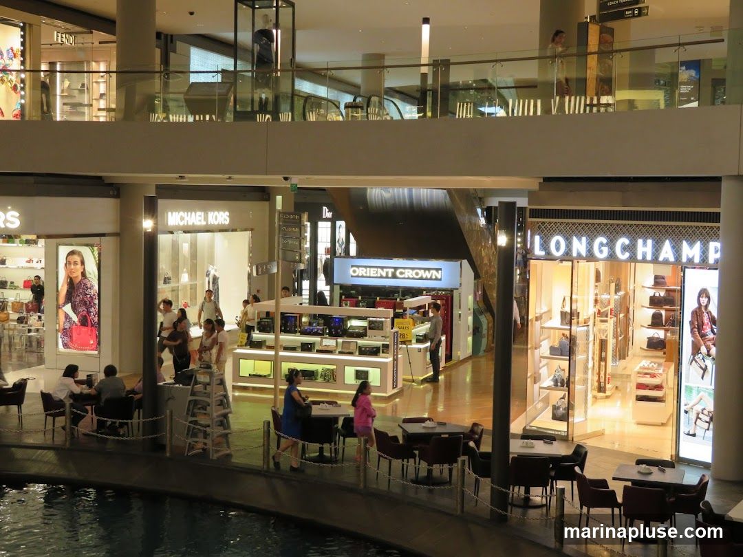 A view of a multi-level shopping mall interior. On the ground level, several storefronts are visible, including Michael Kors and Longchamp. People are seen browsing and sitting at tables in a cafe area. The upper level features a walkway with more shops and escalators.