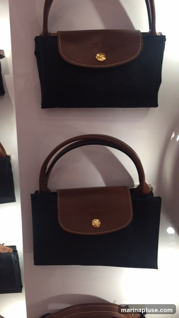 A close-up, vertical shot showcases several black nylon handbags with brown leather trim and gold-tone clasps, arranged on a white display shelf. The bags have a classic, structured design with top handles. The lighting highlights the textures of the leather and the sheen of the metal hardware.