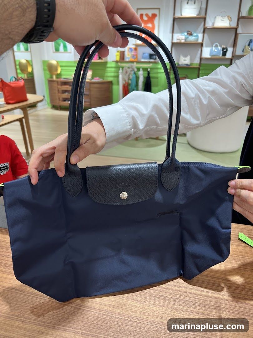 A person holds a navy blue tote bag with black leather trim and handles. The bag has a flap closure with a silver snap. In the background, a retail store is visible with shelves displaying handbags and clothing racks with garments.