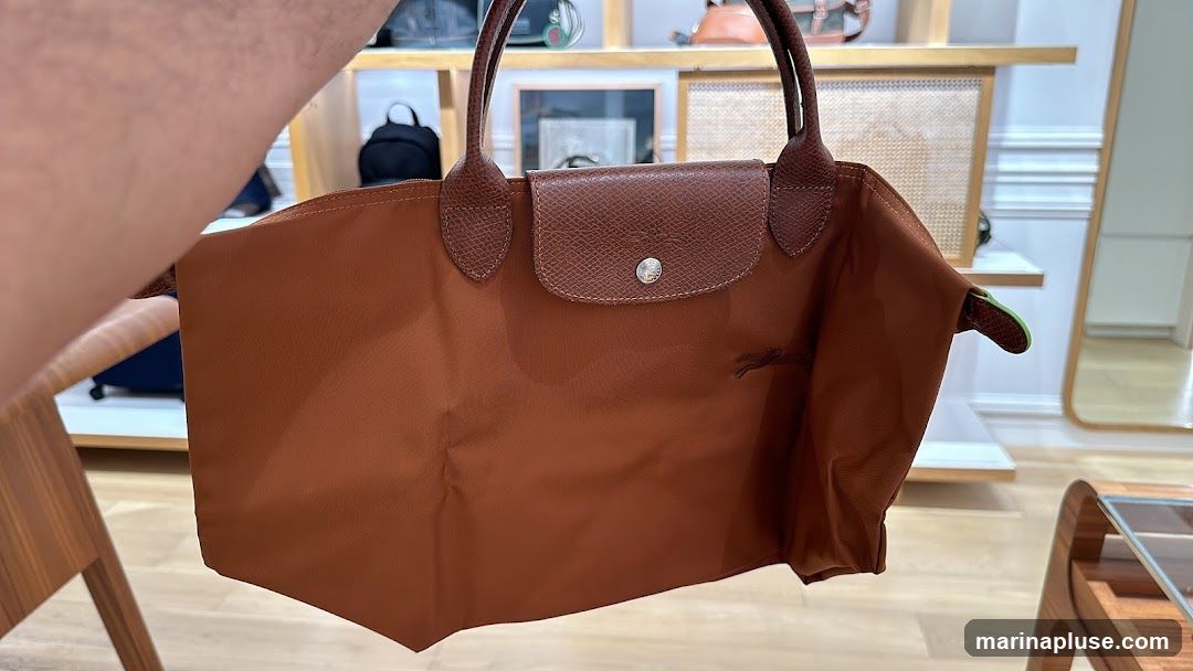 A person is holding a brown nylon tote bag with leather trim and handles. The bag has a flap closure with a snap button. The interior of the bag appears to have a green lining. In the background, there are shelves displaying various bags and accessories, as well as a mirror.