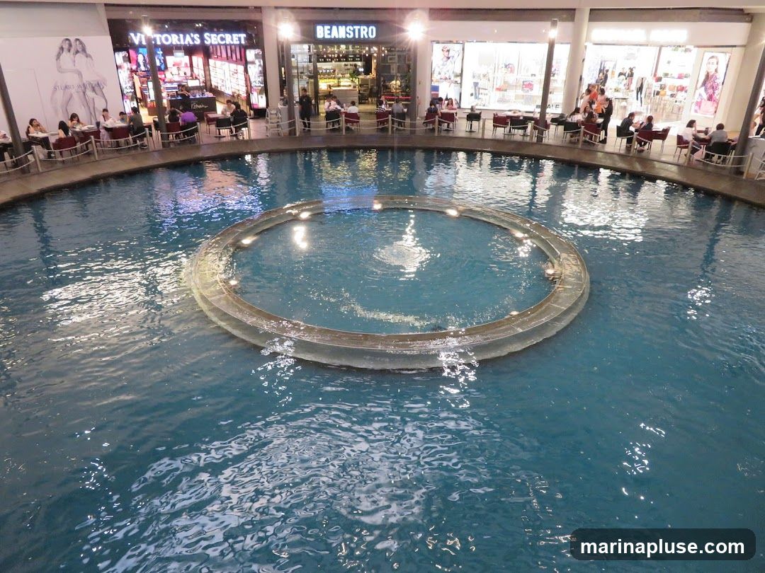 An indoor fountain with a circular structure in the center is surrounded by shops and seating areas. People are seated at tables, some dining and some appearing to be shopping. The shops visible include Victoria's Secret and Beanstro. The overall scene depicts a bustling commercial and recreational space.