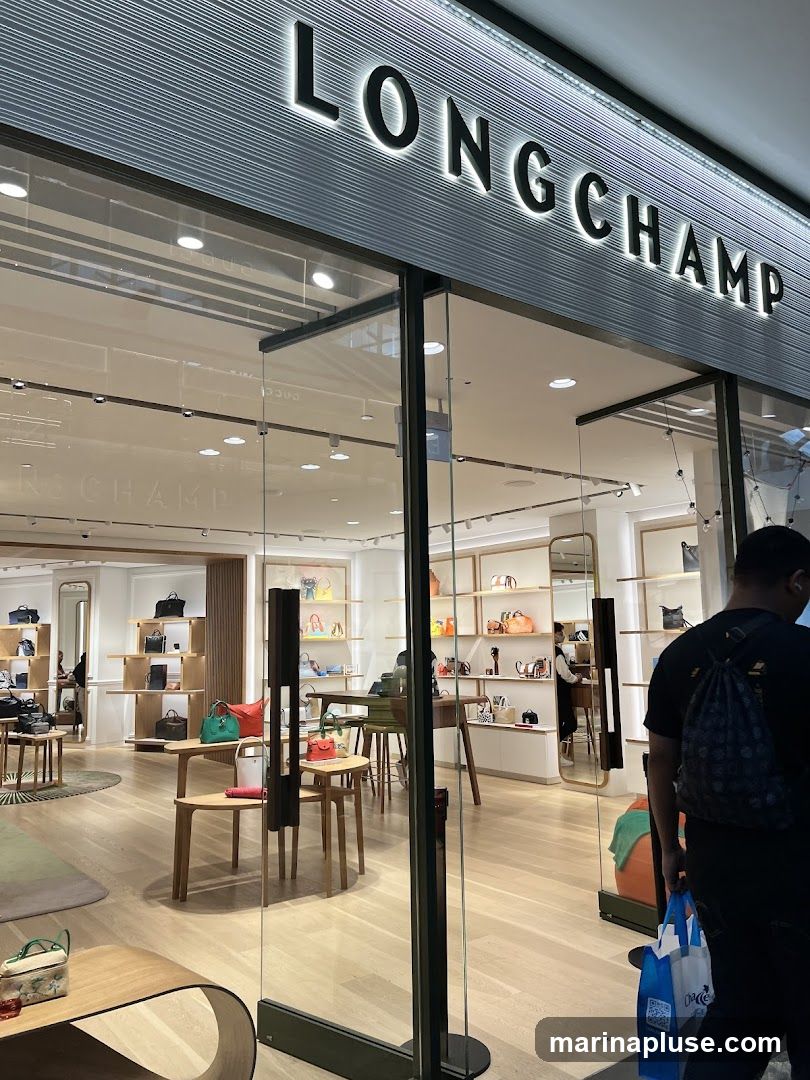 The exterior of a Longchamp store with the brand name illuminated above the entrance. The store features large glass windows showcasing handbags and accessories displayed on shelves and tables inside. A person is seen entering the store carrying shopping bags.
