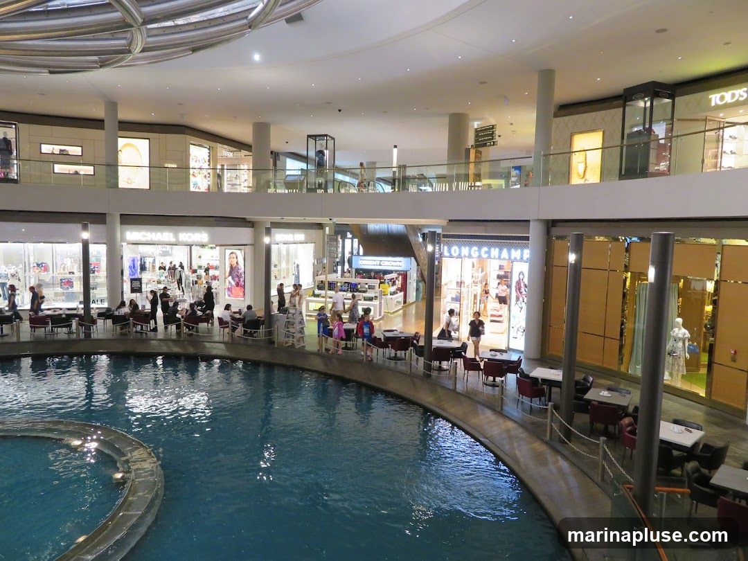 An interior view of a multi-level shopping mall with a large indoor water feature in the foreground. Shops such as Michael Kors and Longchamp are visible on the lower level, with seating areas arranged around the water. The upper levels show more shops and walkways. People are seen browsing and dining.