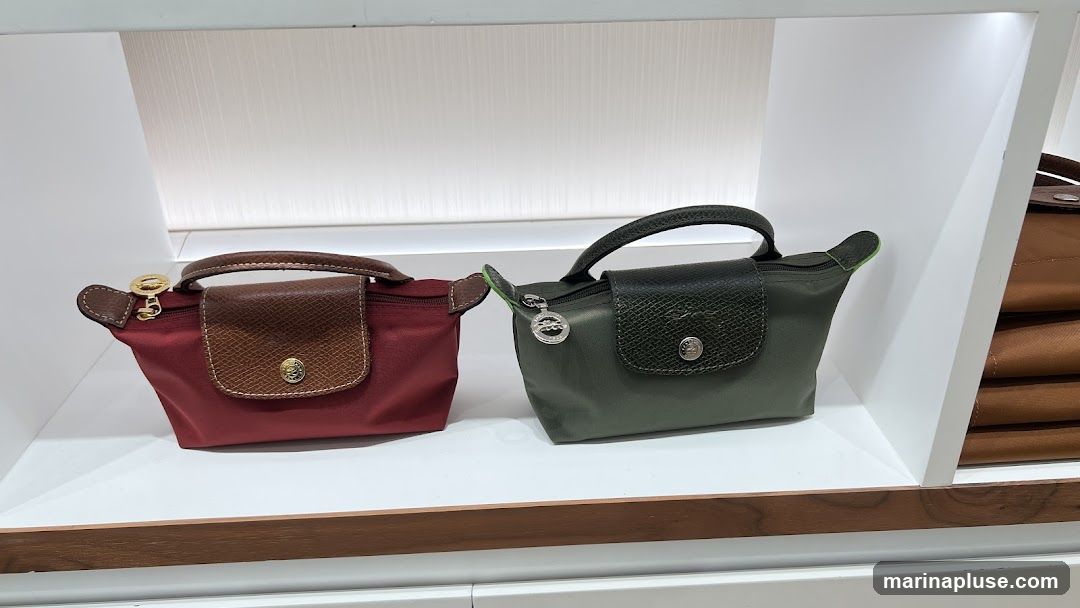 Two small handbags, one red and one green, are displayed on a white shelf. Both have brown leather accents and gold-toned hardware. The red bag has a brown leather flap with a circular emblem. The green bag has a dark green textured leather flap with a similar emblem. To the right, a stack of brown bags is visible.