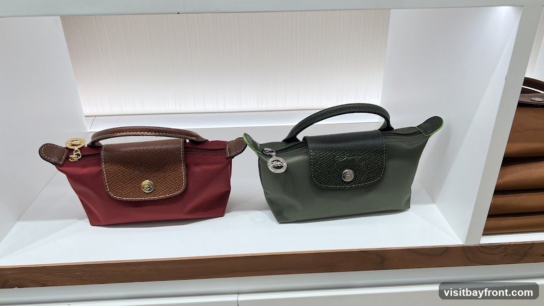 Two small handbags, one red and one green, are displayed on a white shelf. Both have brown leather accents and gold-toned hardware. The red bag has a brown leather flap with a circular emblem. The green bag has a dark green textured leather flap with a similar emblem. To the right, a stack of brown bags is visible.