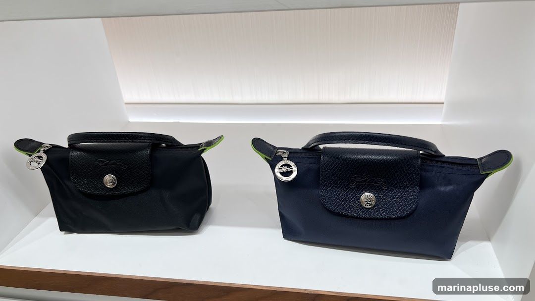 Two Longchamp cosmetic bags are displayed on a white shelf. The bag on the left is black, and the bag on the right is navy blue. Both bags have leather trim and a silver zipper closure. A silver charm with the Longchamp logo hangs from the zipper pull of the navy bag.