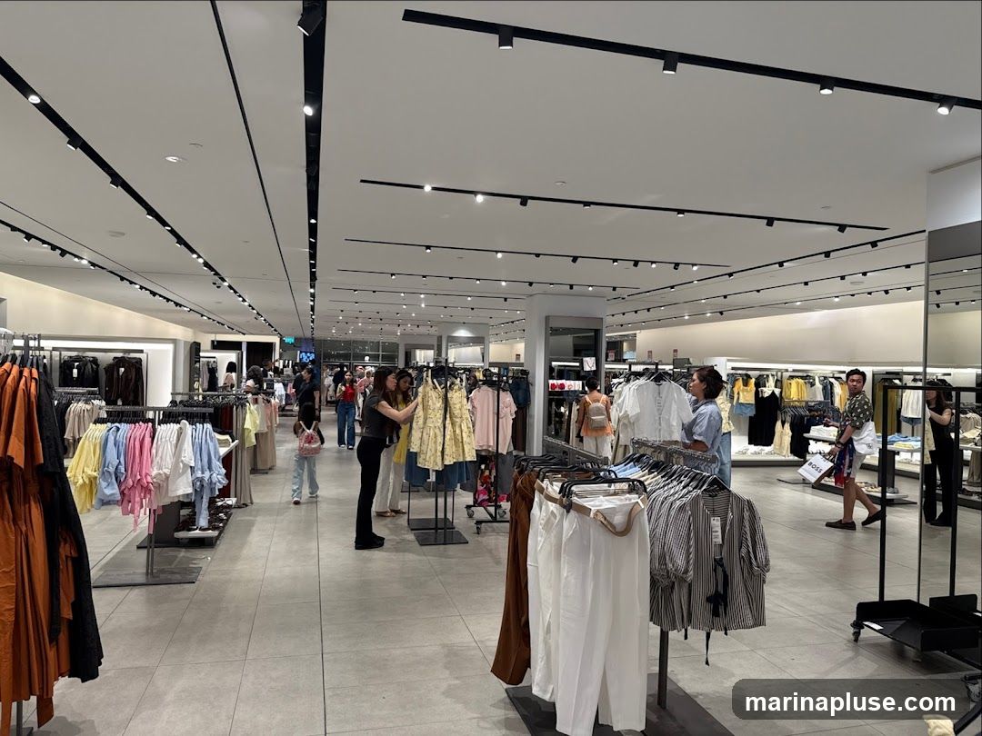 A wide shot of the interior of a brightly lit clothing store. Racks of clothing are displayed on the left and in the center. Several customers are browsing the merchandise. The store has a modern design with a white ceiling and track lighting.