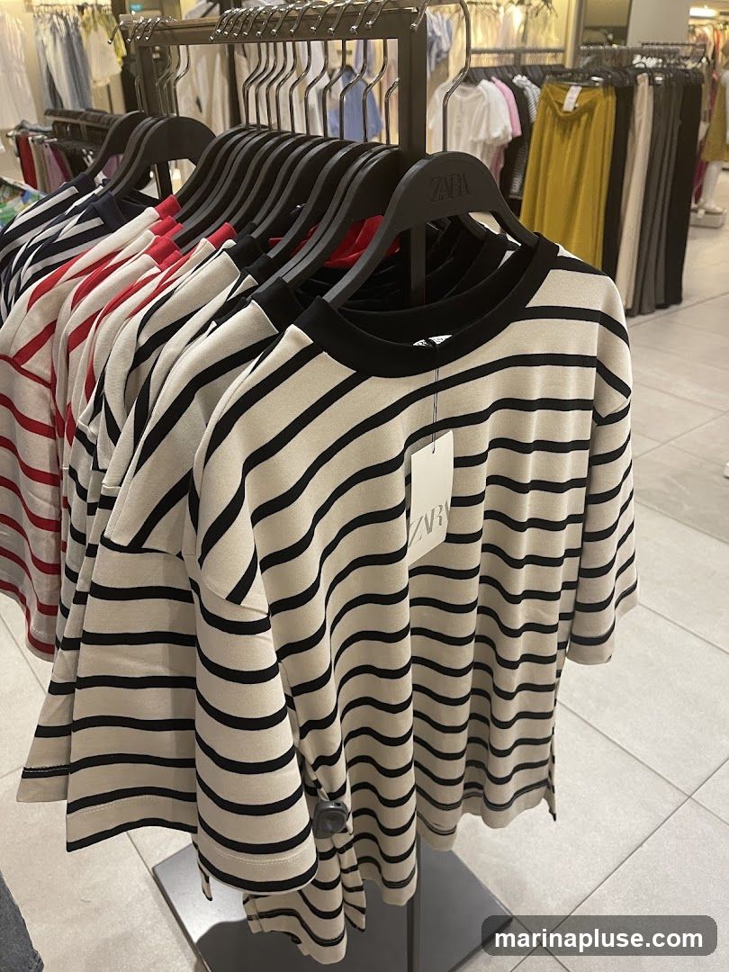 A rack of striped t-shirts in various colors, including red, white, and black, are displayed in a store. The t-shirts are hanging on black hangers. The Zara logo is visible on some of the hangers. In the background, other clothing items and racks are visible, suggesting a retail environment.