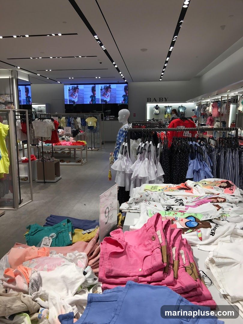 A wide shot of the interior of a clothing store, likely for children, with racks of clothes and displays of folded garments. A large screen at the back shows images of children. The word "BABY" is visible on a sign. There are also price tags and promotional signs visible.