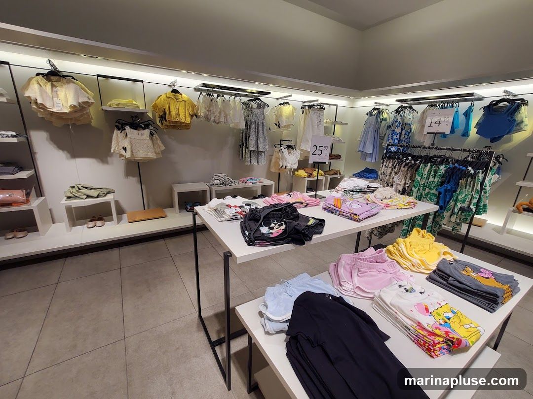 A well-lit interior view of a clothing store, showcasing a variety of apparel for sale. Racks and tables are neatly arranged with folded and hung garments, including dresses, tops, shorts, and t-shirts. Several items feature cartoon characters. A sign indicates "25% OFF" on some items, and another sign displays "14.90 S". The overall impression is a clean and organized retail environment.