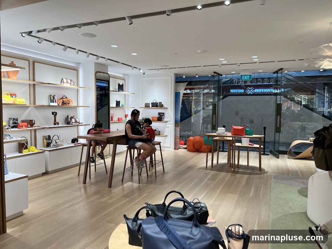The interior of a modern handbag store is shown, with shelves displaying various handbags and accessories. Two children and a woman are seated at a long wooden table in the center of the store. Through the glass doors at the back, an 'EXIT' sign and reflections of other shops are visible. Several large handbags are placed on the floor in the foreground.