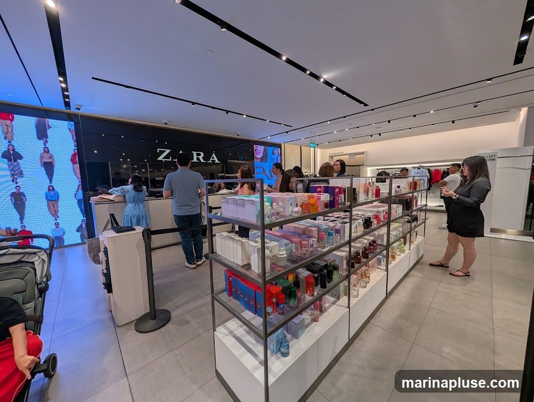 The interior of a Zara store, featuring a display of perfumes and other beauty products on shelves. A large screen on the left displays models. Several customers and staff members are visible in the store.