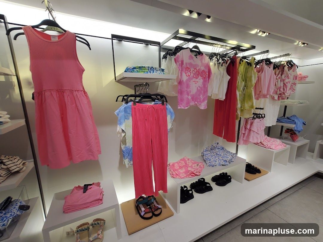 A display of children's clothing in a store. There are pink dresses, pants, shirts, and sandals. The clothes are neatly arranged on shelves and hangers.