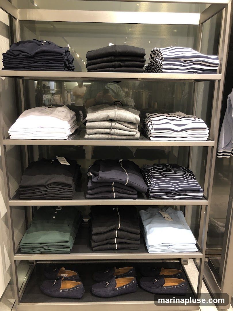 A retail display featuring folded clothing items on shelves and pairs of navy blue loafers on the bottom shelf. The clothing includes t-shirts, polo shirts, and zippered hoodies in various colors such as navy, black, white, grey, green, and light blue. Some items have tags attached. The display is organized on metal shelving units.
