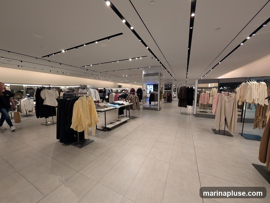 The interior of a well-lit clothing store with racks of apparel displayed on the floor and hanging from racks. There are mirrors and display stands throughout the store. A few customers are visible browsing the merchandise.