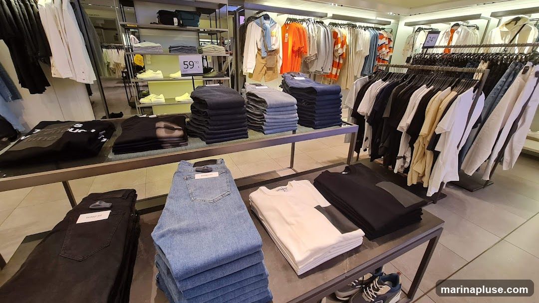A well-lit interior of a clothing store showcasing folded jeans and t-shirts on display tables, with racks of hanging shirts and jackets in the background. A price tag of 59.90 is visible on a shelf with yellow sneakers.