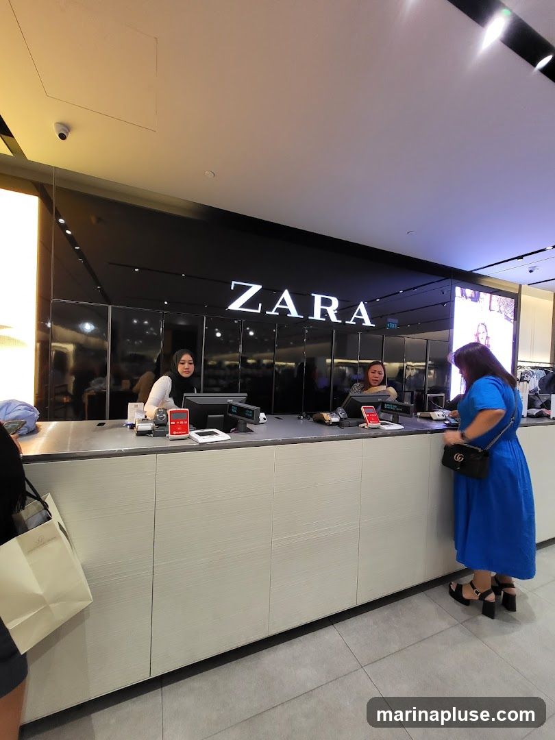 A wide shot of a Zara store's checkout counter. The prominent "ZARA" logo is displayed in large white letters on a black background above the counter. Two employees are visible behind the counter, one wearing a hijab and the other with long hair. A customer in a bright blue dress is standing at the counter, interacting with an employee. There are various items on the counter, including a computer monitor, a credit card reader, and a shopping bag. The counter itself is light-colored with a textured finish.