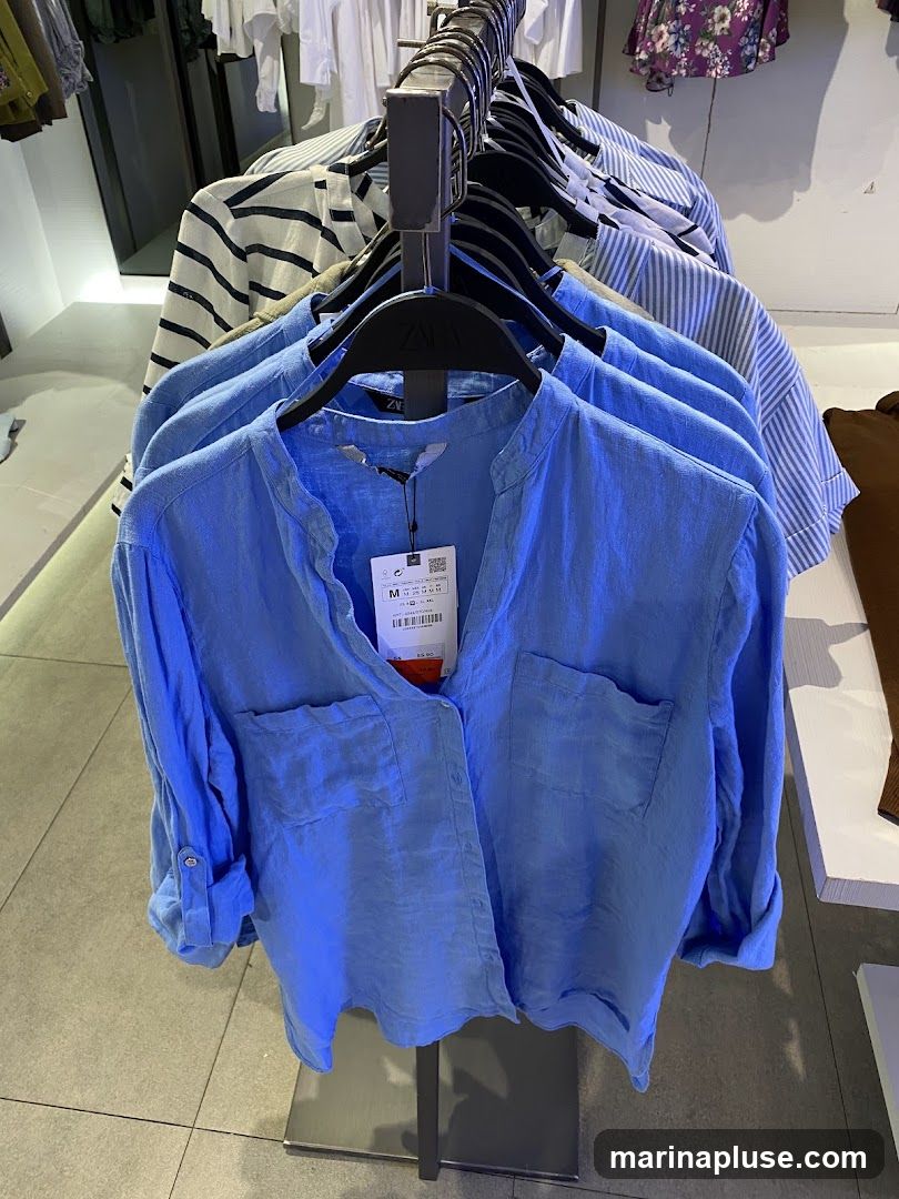 A rack of blue linen shirts is displayed in a store. The shirts have a V-neck and two chest pockets. One shirt has a tag with size information and a price of £25.99. Other shirts on the rack include striped patterns. In the background, more clothing items are visible on racks and shelves.