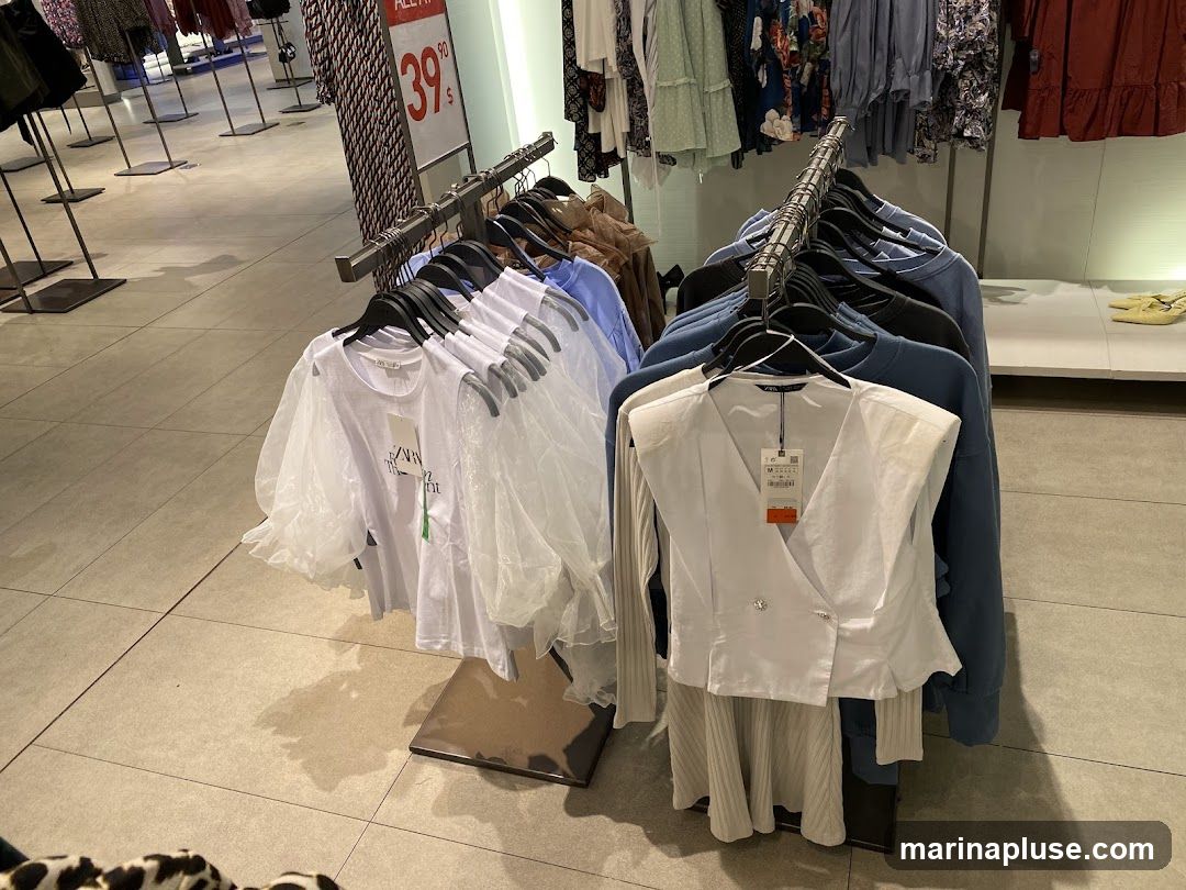 A rack of women's clothing is displayed in a store. The clothing includes white tops with sheer sleeves, a white vest, and blue sweaters. A sign in the background indicates a sale price of 39.