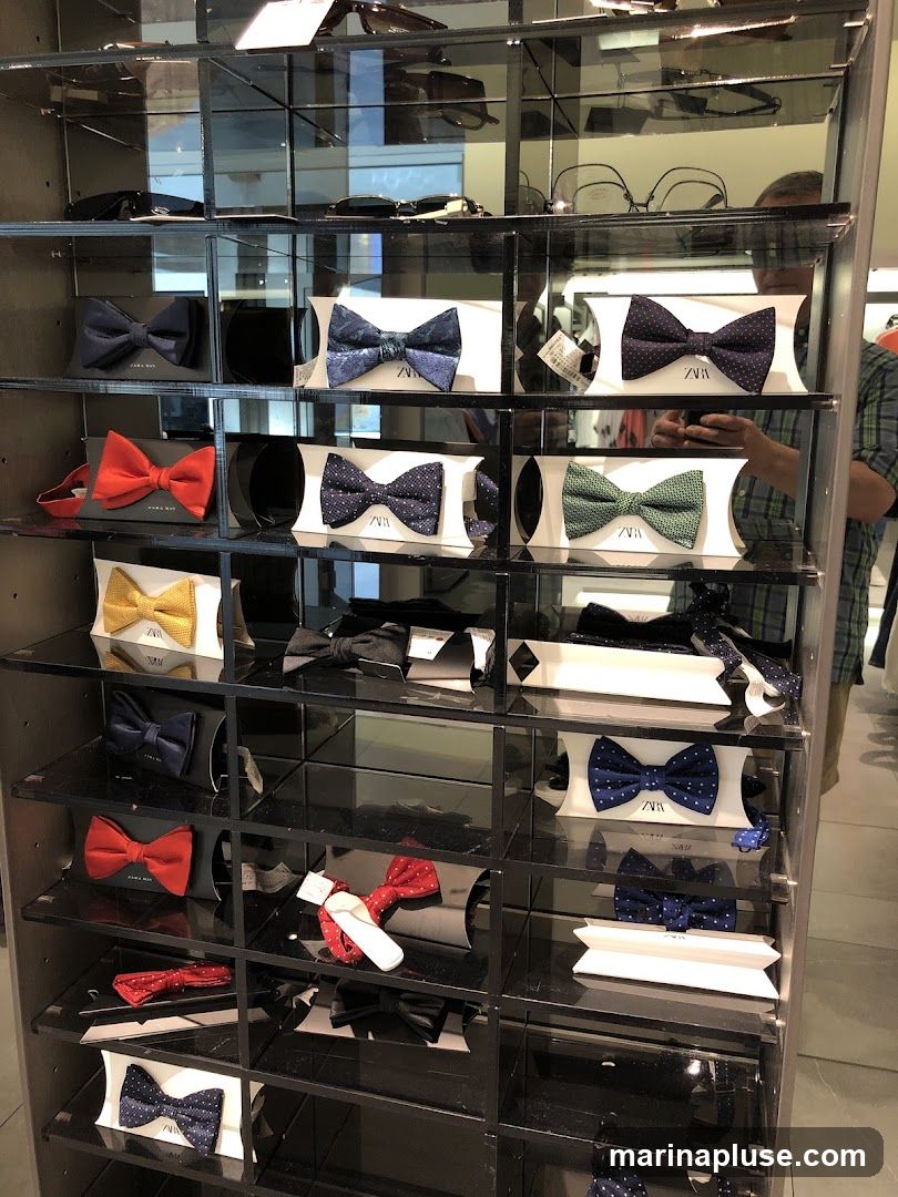 A display case filled with various bow ties. The bow ties are neatly arranged on shelves, some in packaging with the Zara Man logo. Different colors and patterns are visible, including solid navy, red, yellow, and patterned blue and green bow ties. Sunglasses are displayed on shelves above the bow ties.