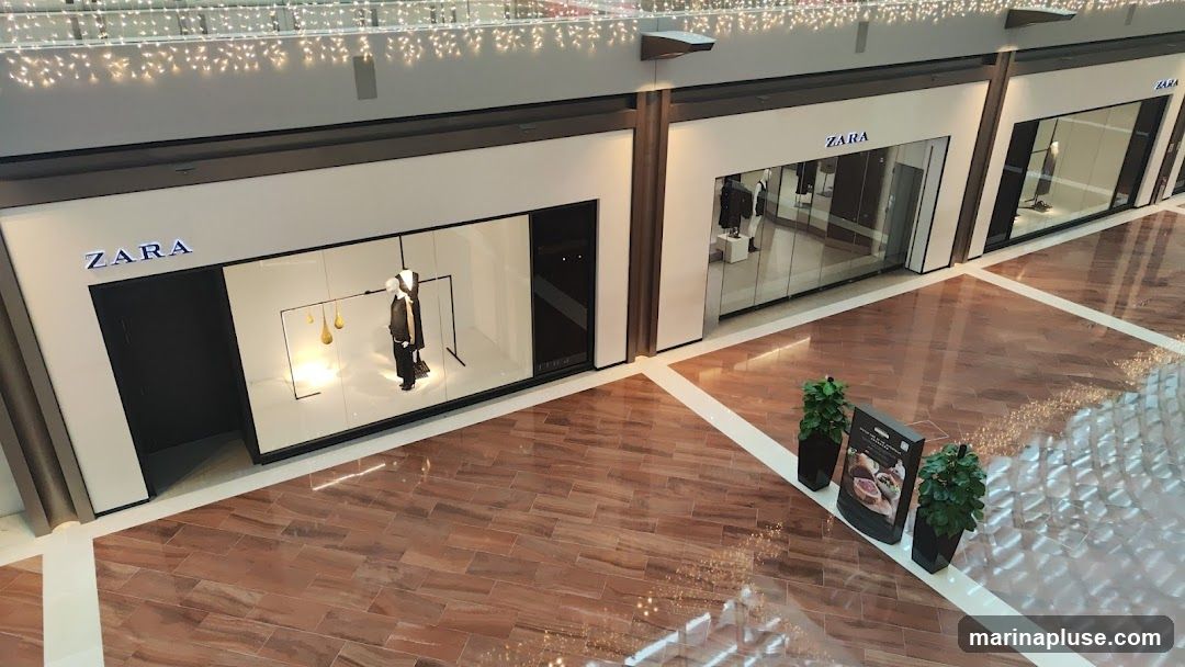 An aerial view of multiple Zara storefronts in a modern shopping mall. The stores have large glass windows displaying mannequins in fashionable clothing. The Zara logo is prominently displayed above each entrance. The mall features polished wooden floors and decorative lighting.