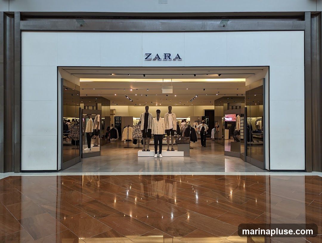The exterior of a Zara clothing store with its name prominently displayed above the entrance. The store features large glass doors and windows, revealing mannequins dressed in fashionable clothing and racks of apparel inside. The interior is well-lit, showcasing a modern retail environment.