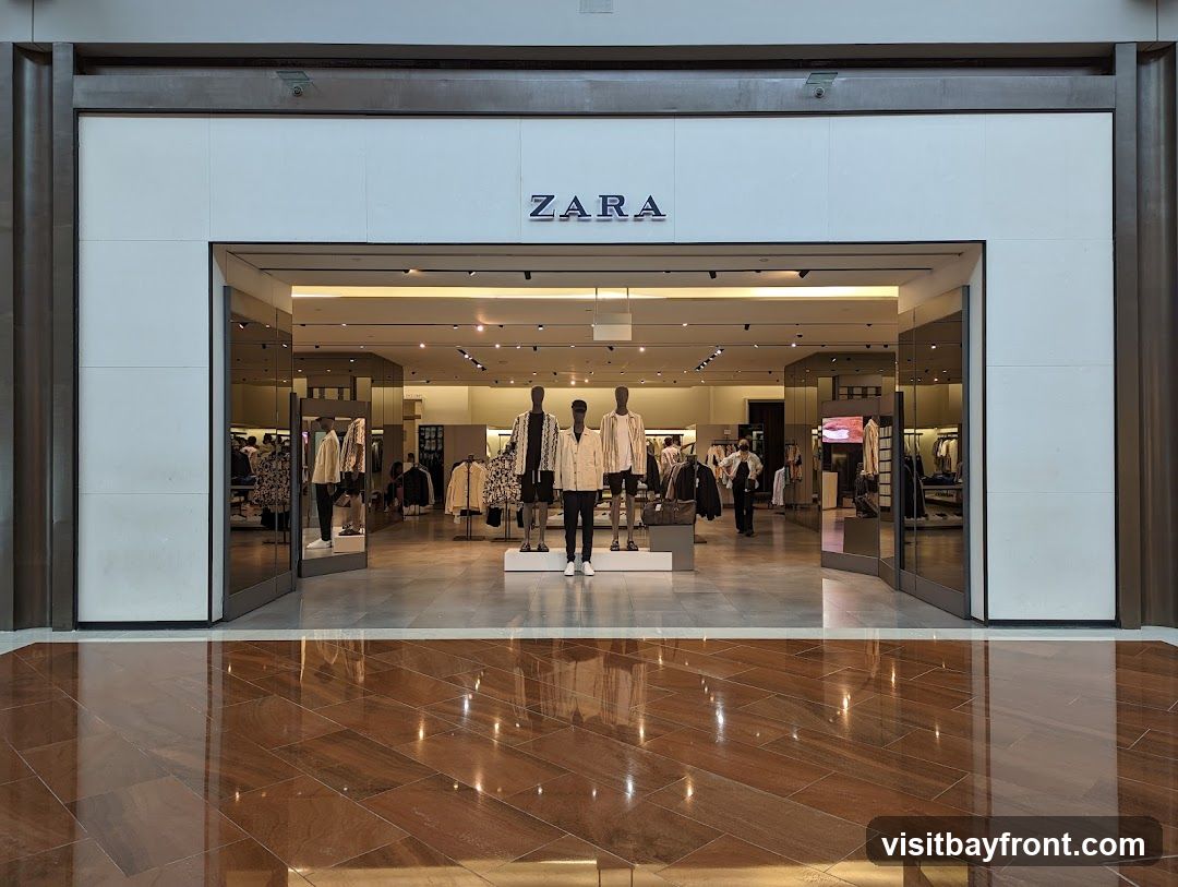The exterior of a Zara clothing store with its name prominently displayed above the entrance. The store features large glass doors and windows, revealing mannequins dressed in fashionable clothing and racks of apparel inside. The interior is well-lit, showcasing a modern retail environment.